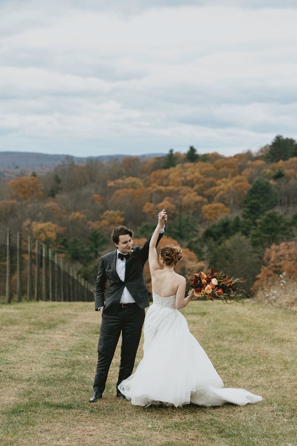 A bride and groom dance playfully in a grassy meadow with autumn mountains and trees in the background.