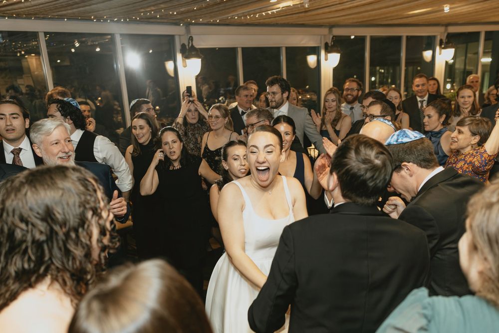 Wedding guests dance and celebrate together on a crowded dance floor under dimly lit reception lighting.