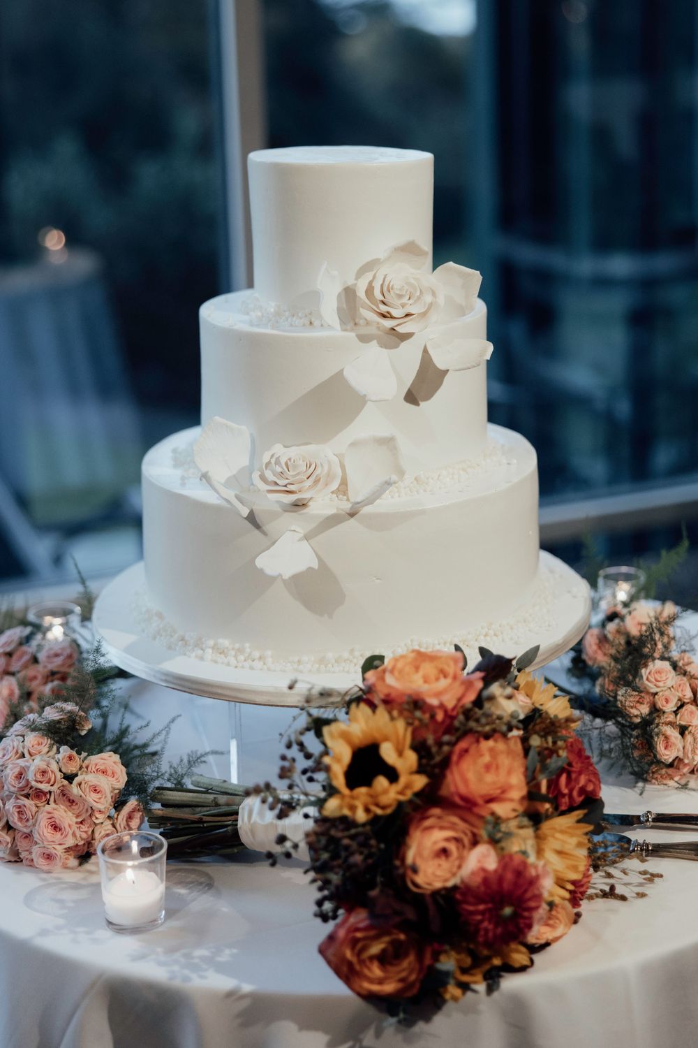 Elegant white wedding cake adorned with delicate roses and surrounded by autumn-toned floral arrangements on display table.