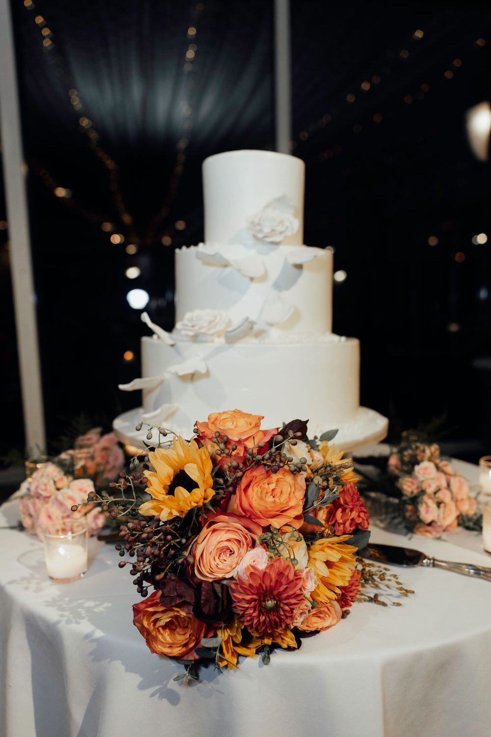 An elegant white wedding cake adorned with autumn-colored flowers and sunflowers on a decorated table.