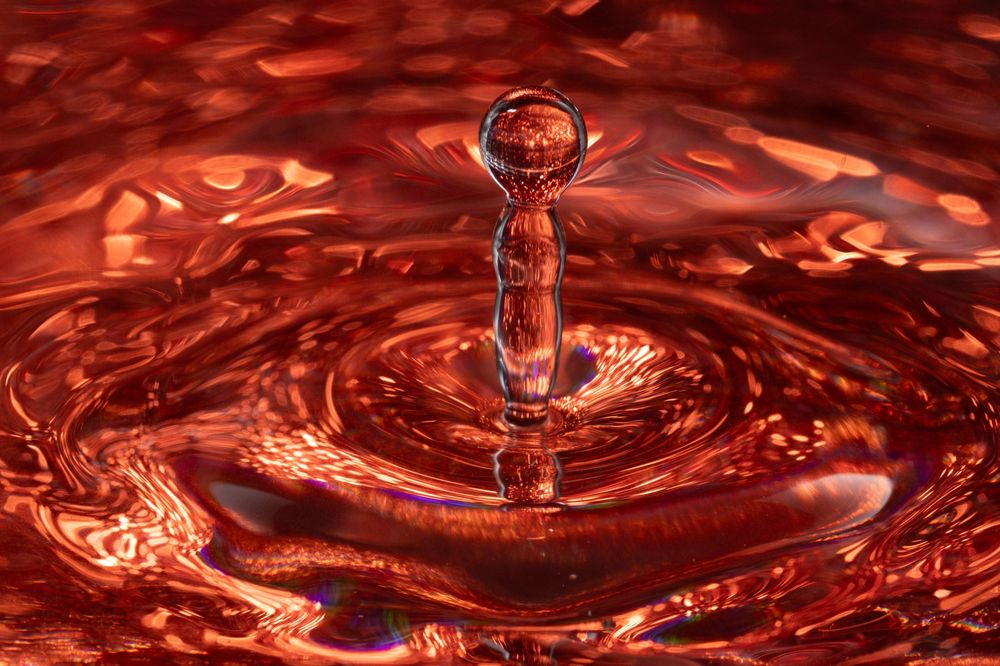 Macro photography captures a series of red water droplets creating ripples and crown-like splashes in liquid.