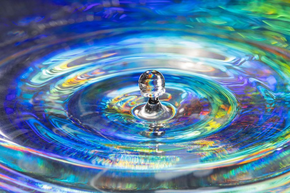 Macro photograph of a water drop creating concentric ripples with iridescent colors.