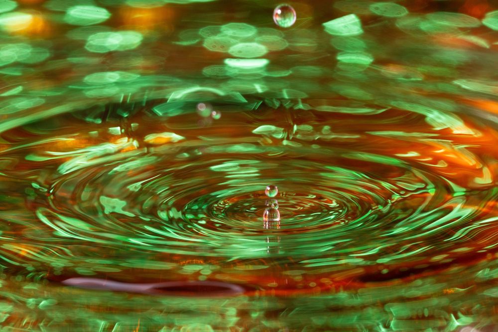 Liquid surface with green and orange reflections showing circular ripple patterns.