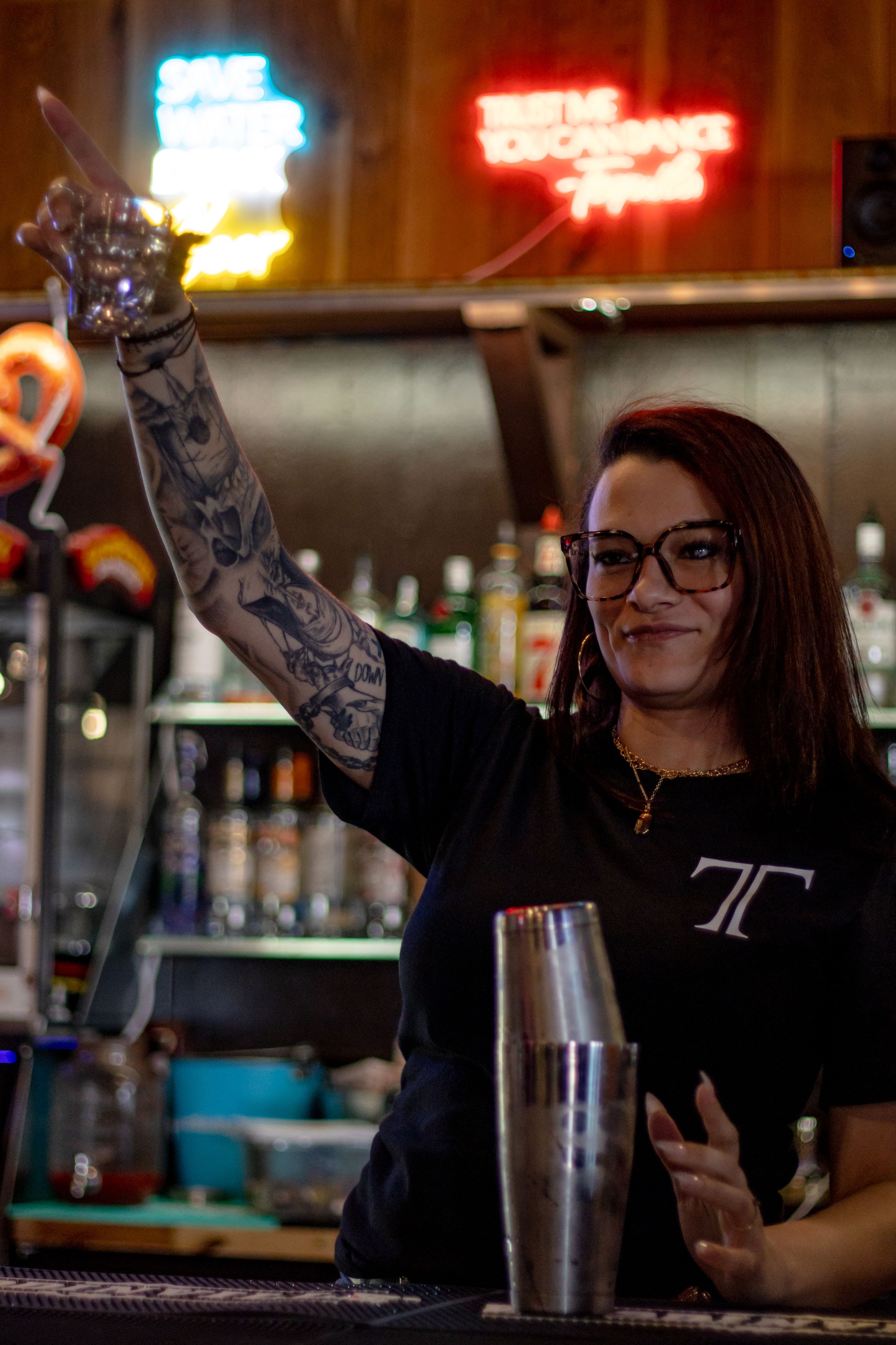 Tattooed female bartender smiling and pointing up behind bar with cocktail shaker at Tommy's Tavern.