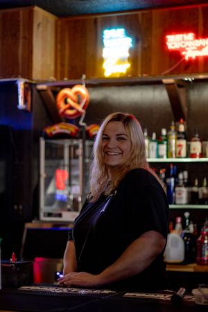 Smiling blonde female bartender standing behind bar with neon signs and liquor bottles.