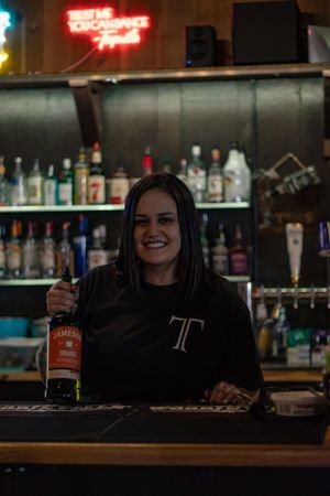 Smiling bartender holding a whiskey bottle behind a well-stocked bar with neon signs at Tommy's Tavern during Yelm marketing photoshoot