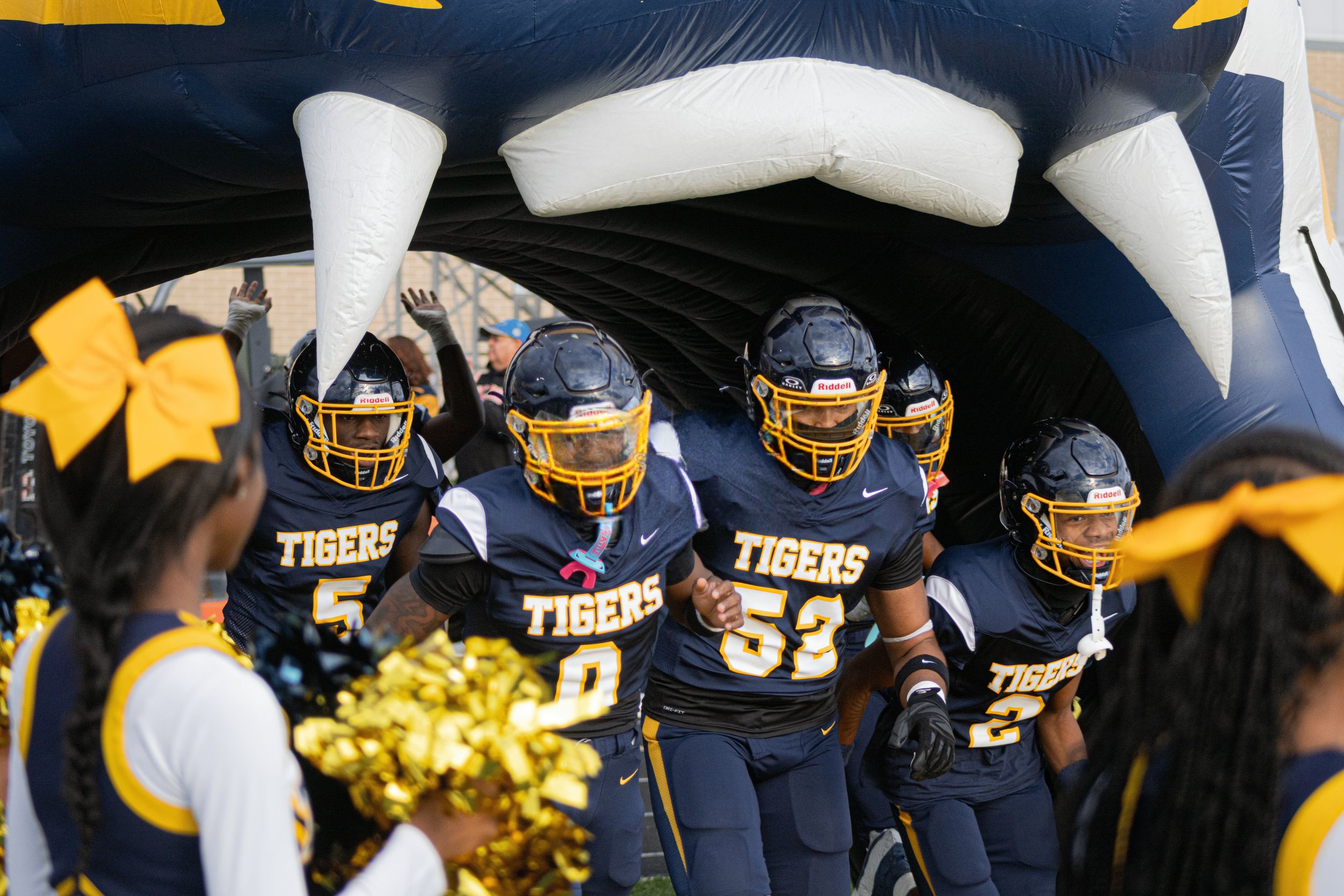 Tigers football players run through an inflatable tunnel onto the field, cheered on by cheerleaders with gold pom-poms.