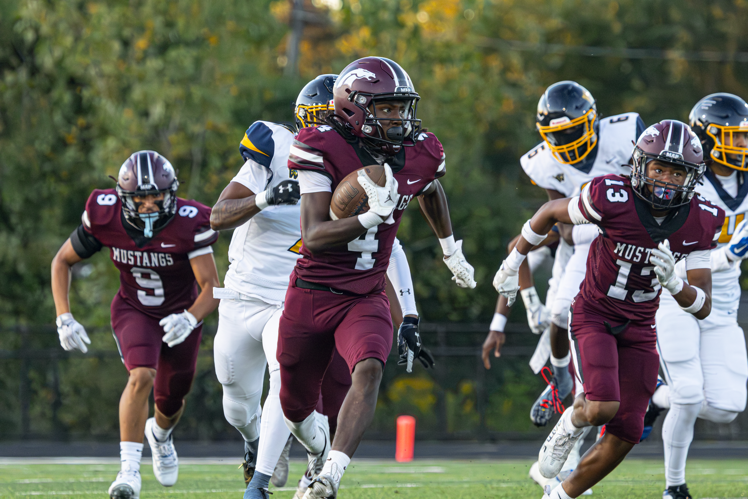 Football player in a maroon Mustangs uniform sprinting with the ball on an outdoor field during a game.