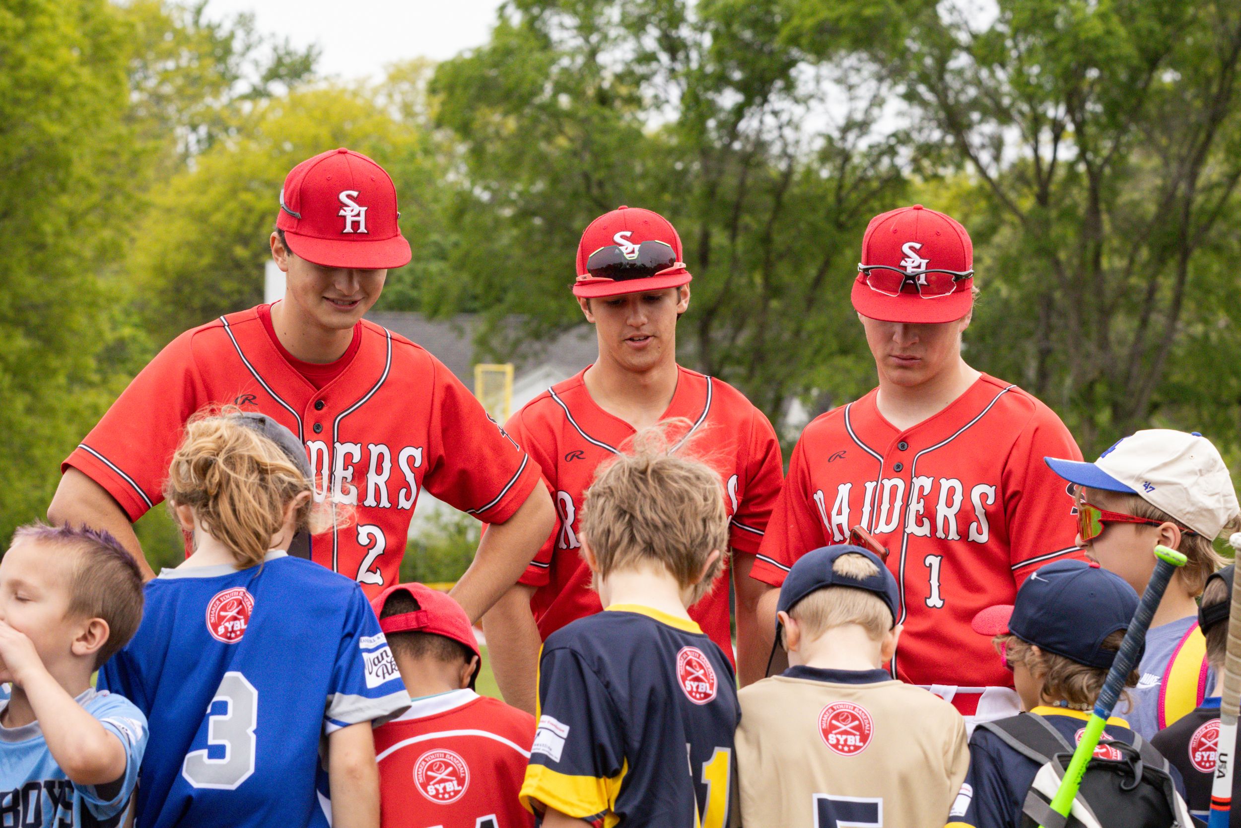 Three Raiders baseball players in red uniforms signing autographs for young fans at an outdoor youth baseball event.