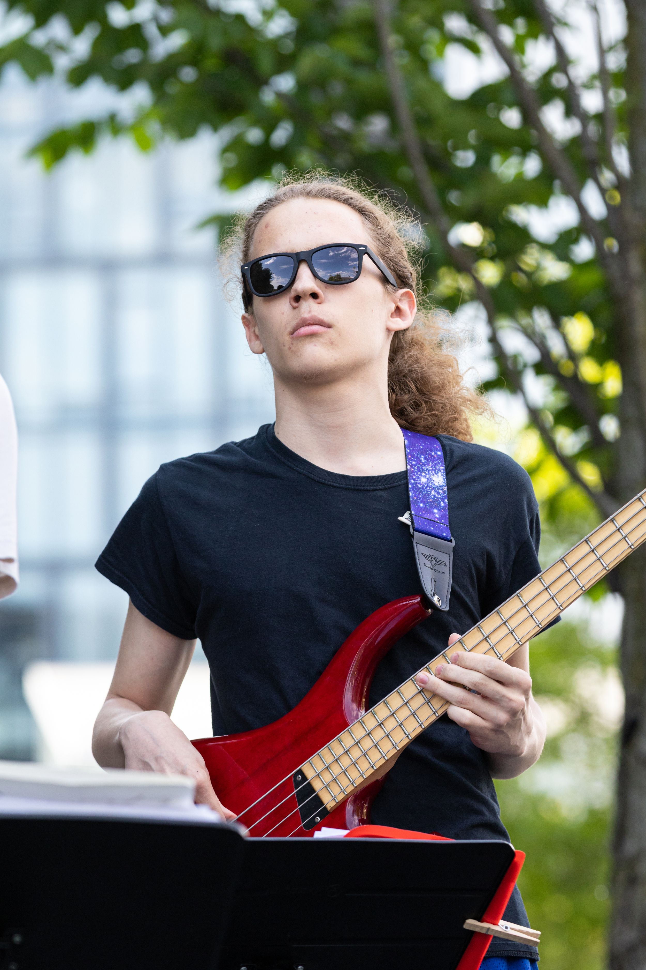 Young musician with curly hair and sunglasses playing a red electric guitar outdoors near trees.