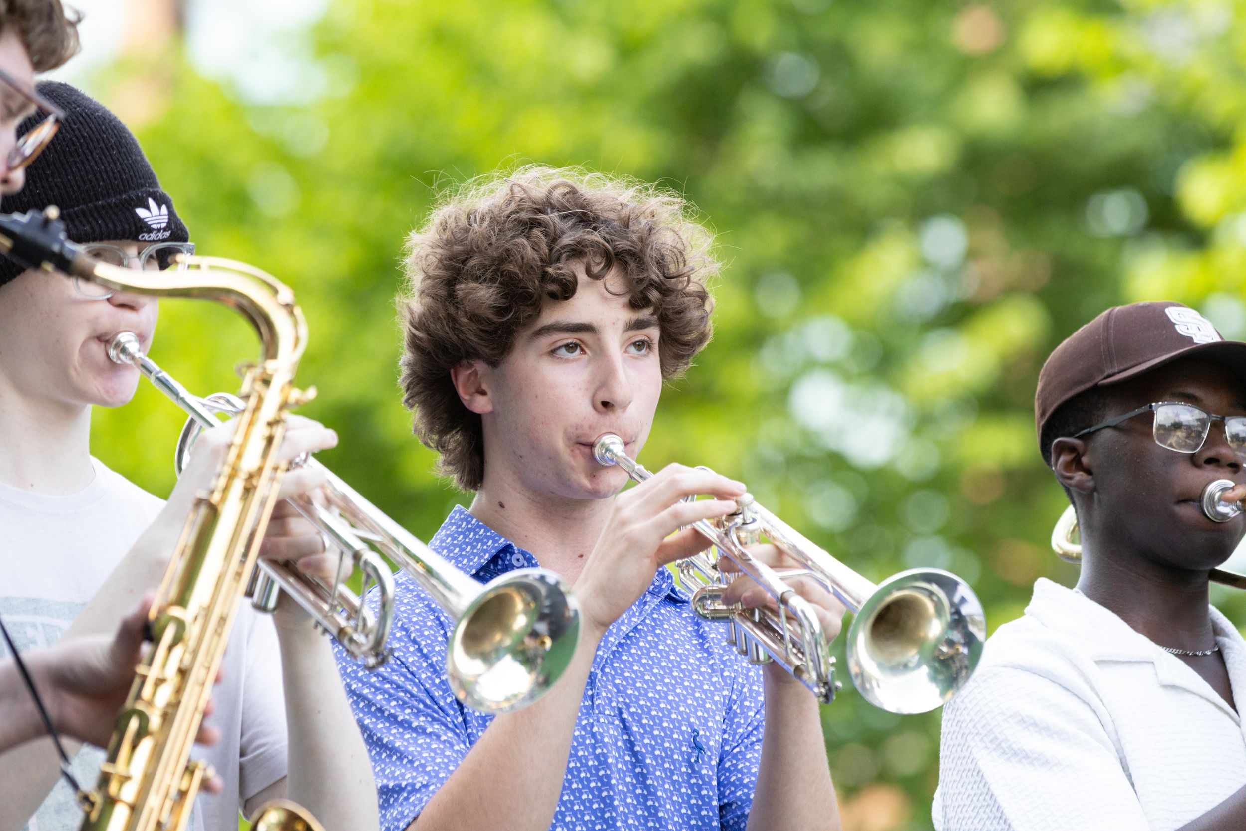 Young musicians playing trumpet and saxophone outdoors at a summer music event surrounded by green trees.