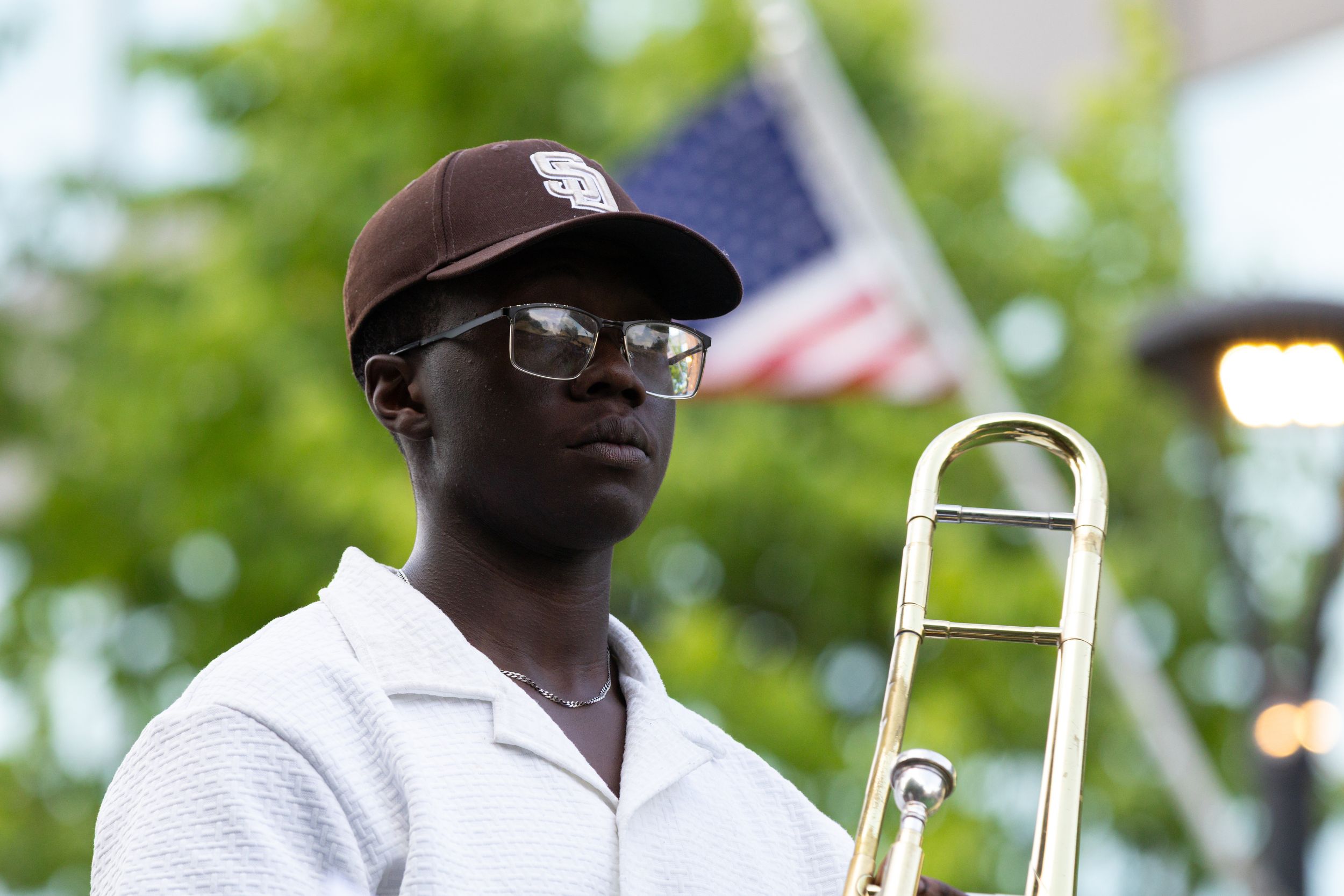 Young musician holding a trombone outdoors near an American flag at a public event.