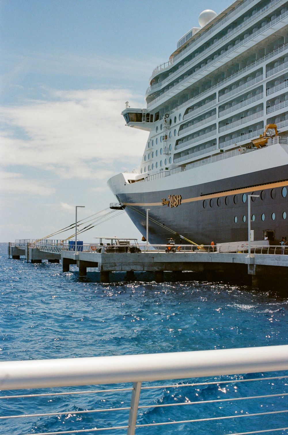 Large cruise ship docked at a pier on calm blue ocean waters under a partly cloudy sky.