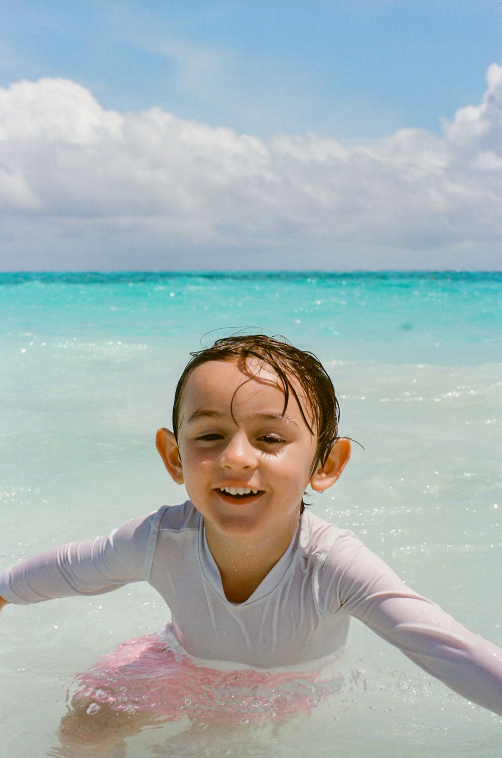 A happy child splashing in crystal-clear turquoise ocean water on a sunny beach day wearing a white rash guard.