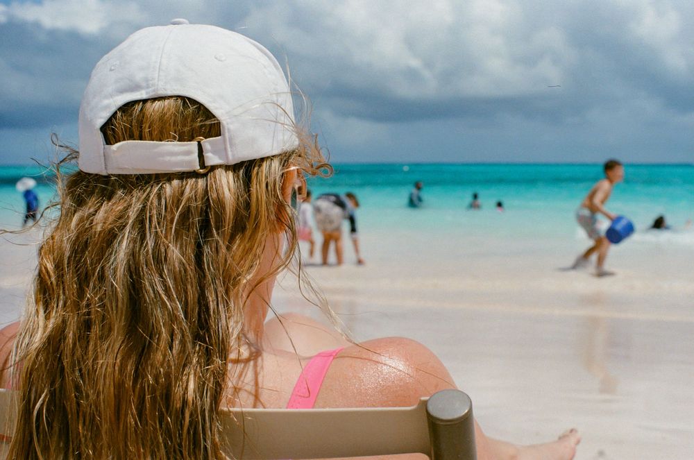 Woman in white cap and pink bikini sits on beach chair overlooking turquoise ocean with people playing in the water.