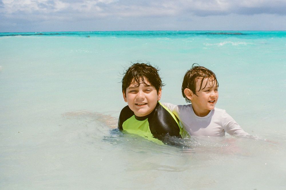 Two children playing in clear turquoise ocean water, one wearing a green life vest, smiling at a tropical beach.