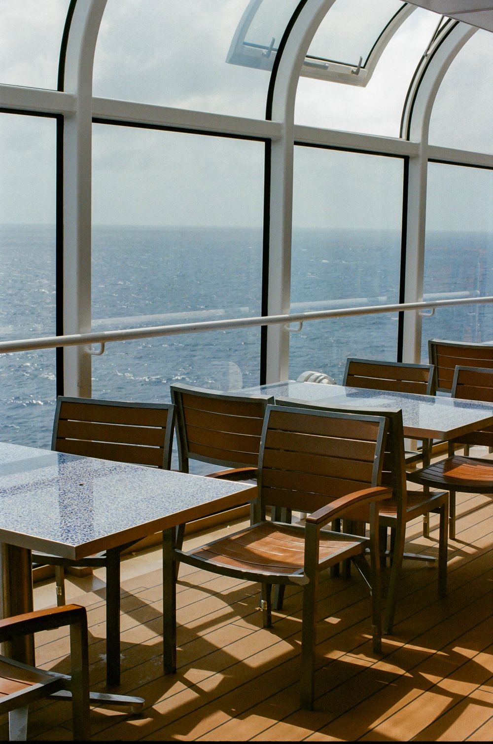 Sunny cruise ship dining area with wooden chairs, marble tables, and panoramic ocean views through curved glass windows.