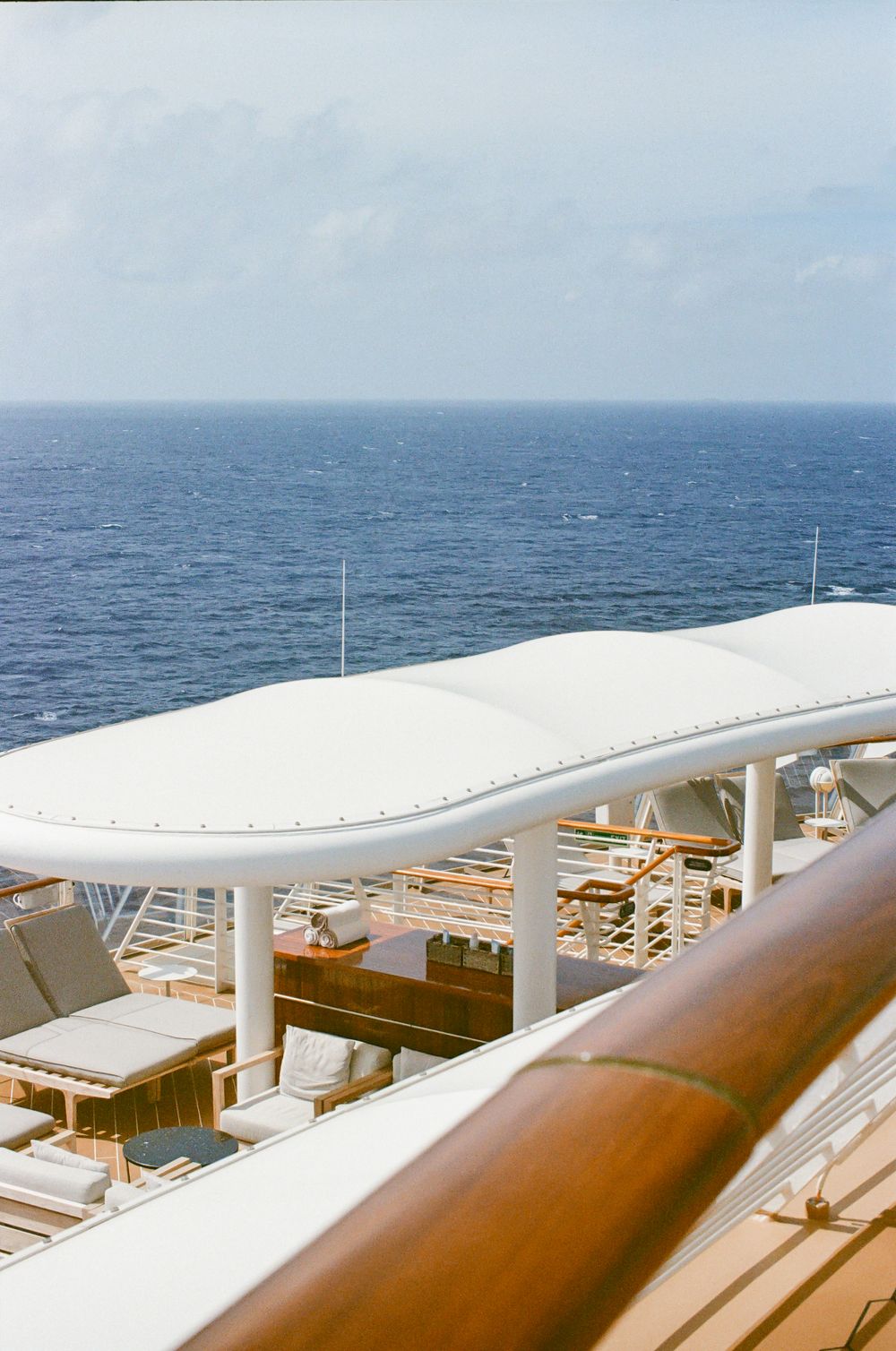 Cruise ship upper deck with white canopy, lounge chairs, wooden railing, and vast ocean horizon under cloudy sky.