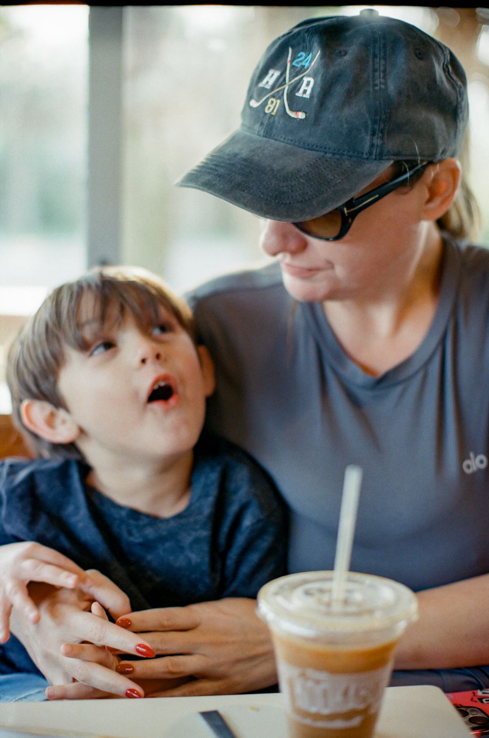 A woman in a baseball cap smiles at a young boy at a cafe table with an iced coffee nearby.