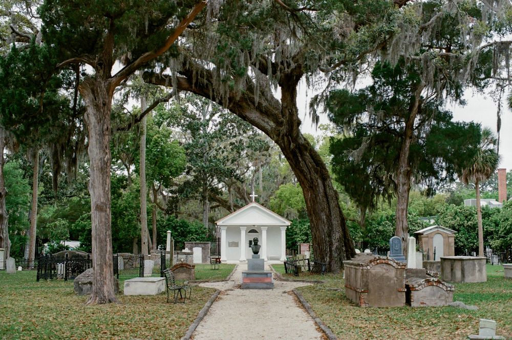 Historic cemetery with moss-draped oak trees framing a white chapel mausoleum surrounded by aged gravestones.