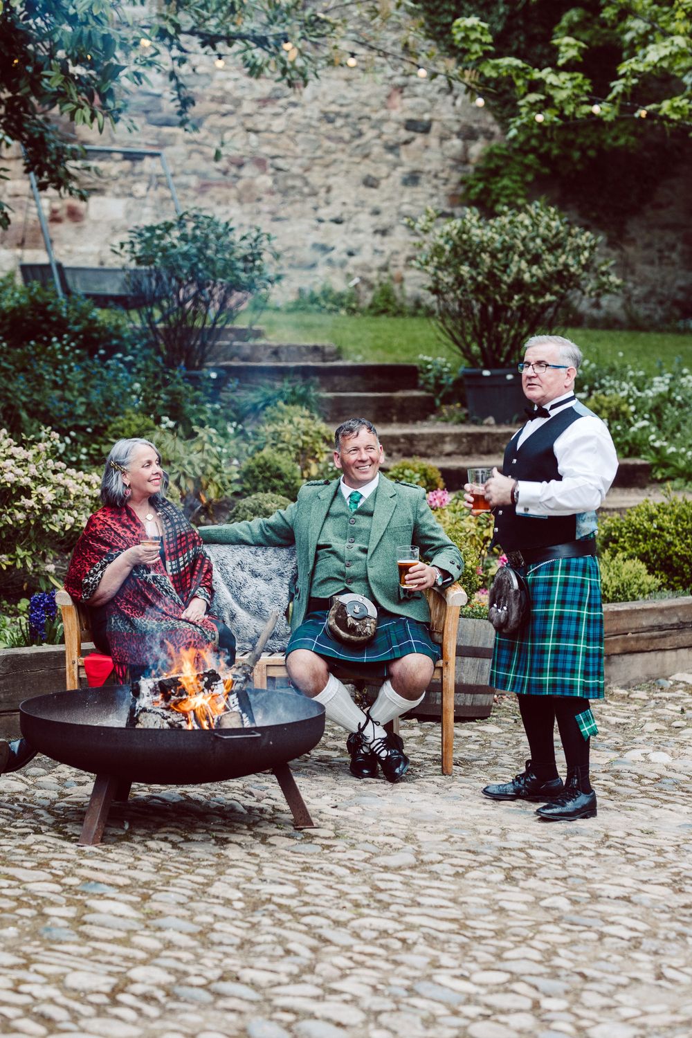 A group of people sitting around an outdoor fire pit on a cobblestone patio enjoying drinks and conversation.