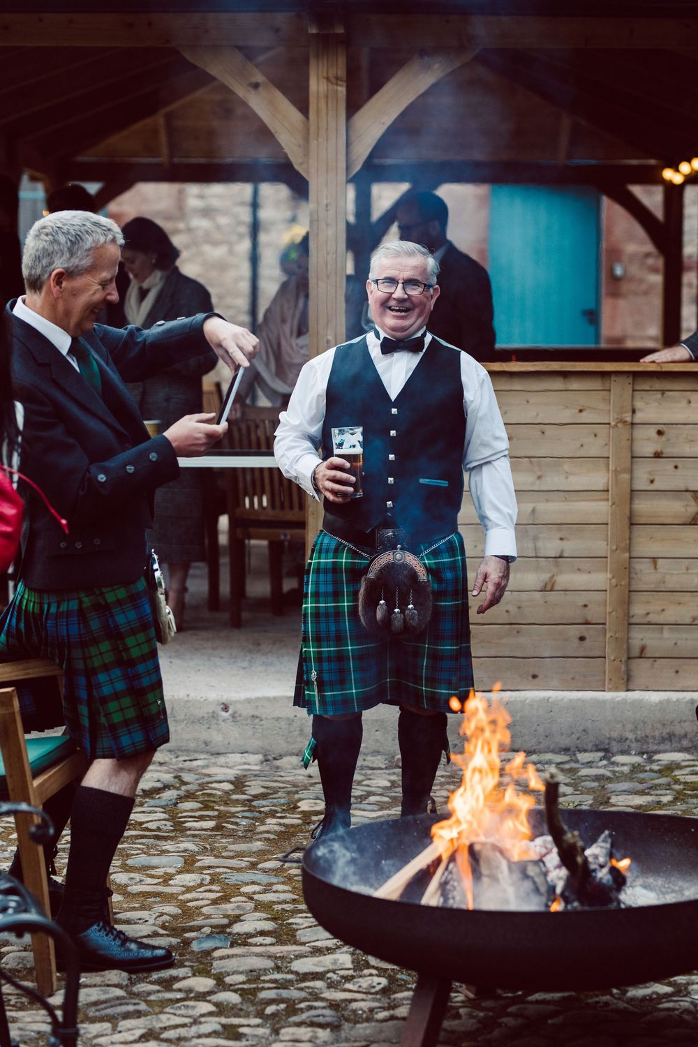 People in traditional Scottish kilts gather around a fire pit during an outdoor celebration event.