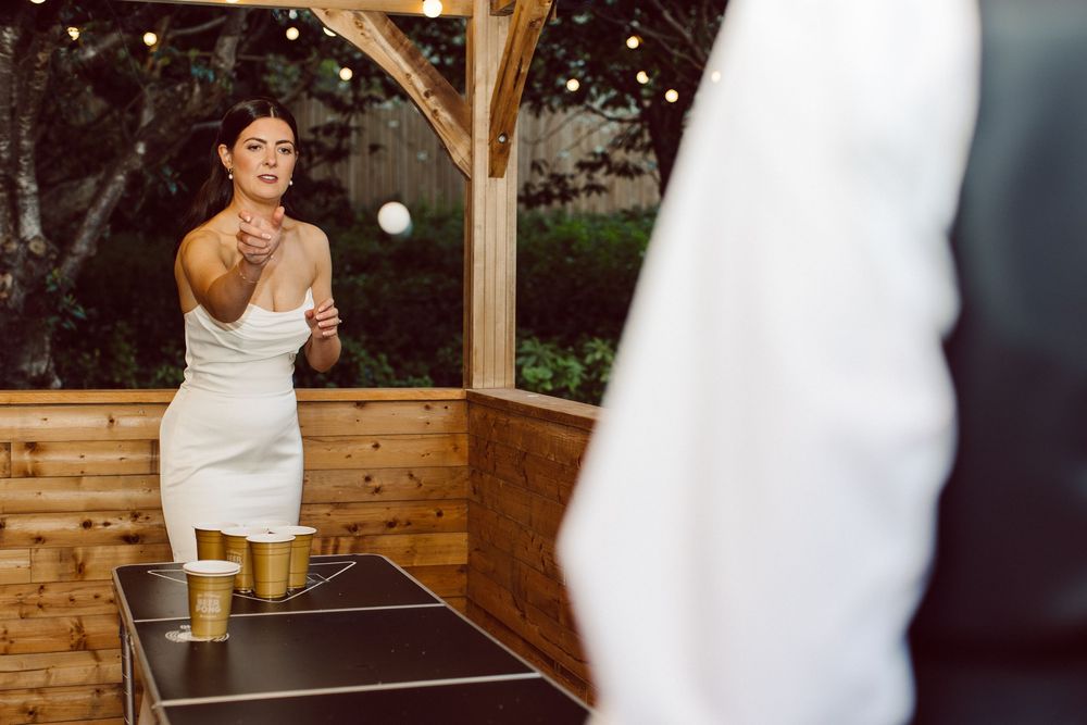 Person in white dress playing ping pong at outdoor party venue under wooden gazebo with beer on table.
