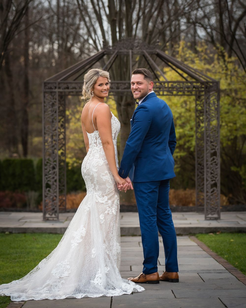 A couple in wedding attire poses together on a garden path with ornate metal archways in the background.