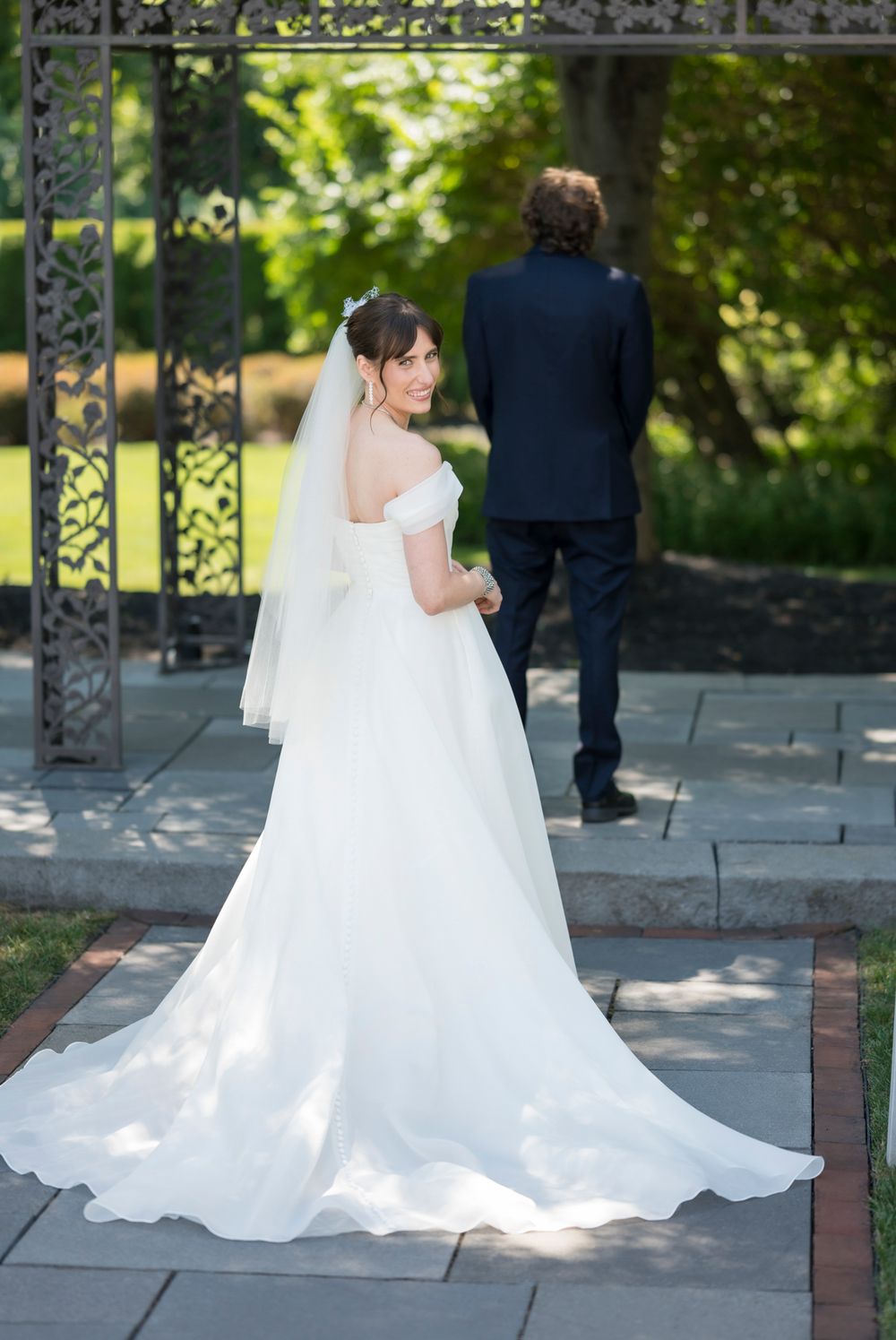 Elegant white wedding dress with flowing train on stone patio surrounded by ornate iron gates and greenery.