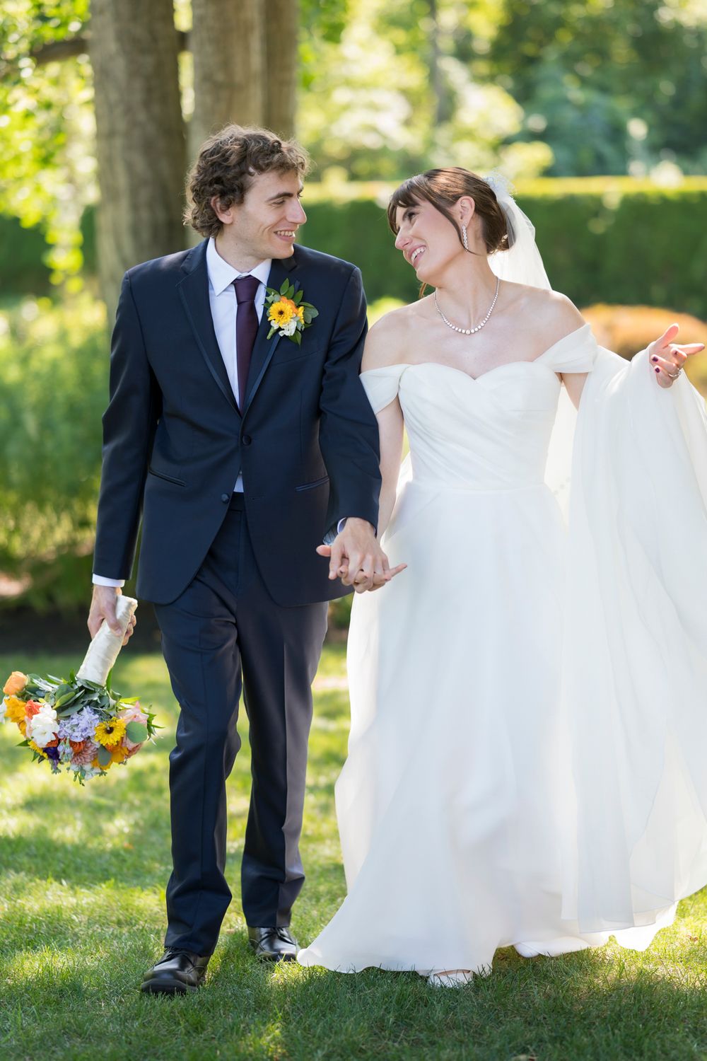 A couple in wedding attire shares a joyful moment together on a sunny lawn surrounded by trees.