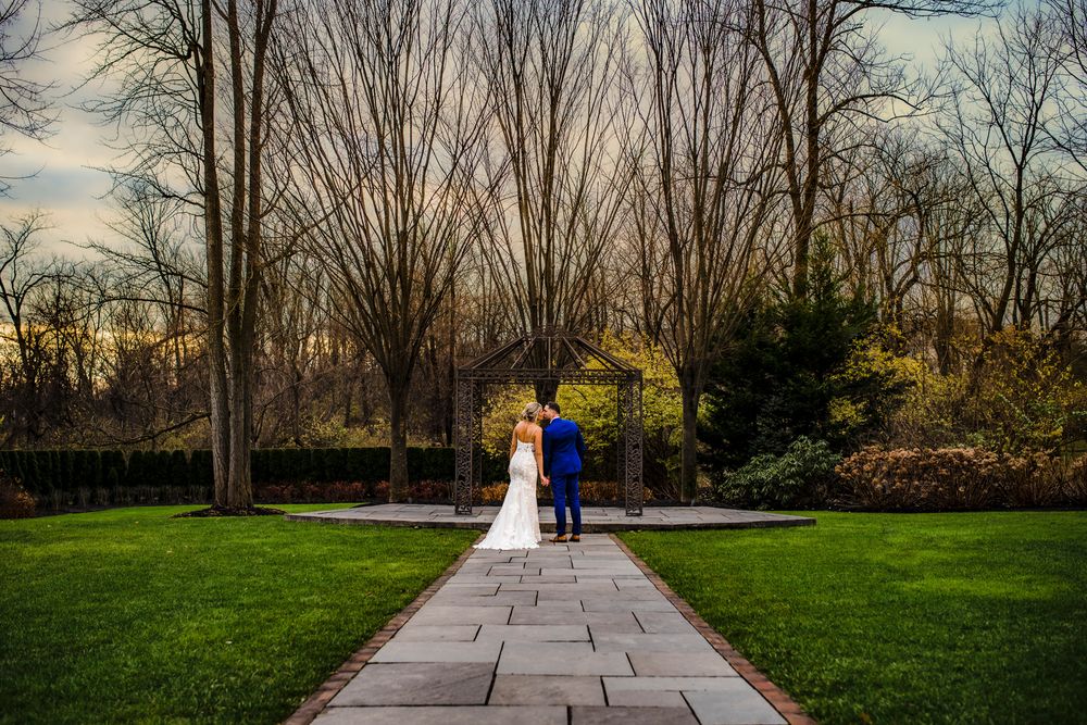 A couple in wedding attire walks down a stone pathway surrounded by lush green lawns and bare winter trees at sunset.