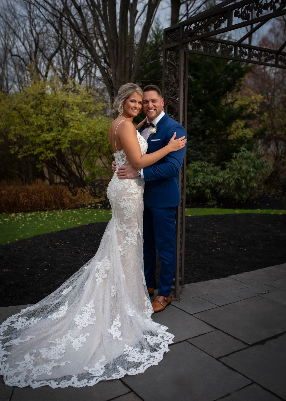 A couple in wedding attire dance together on a stone patio surrounded by autumn trees.