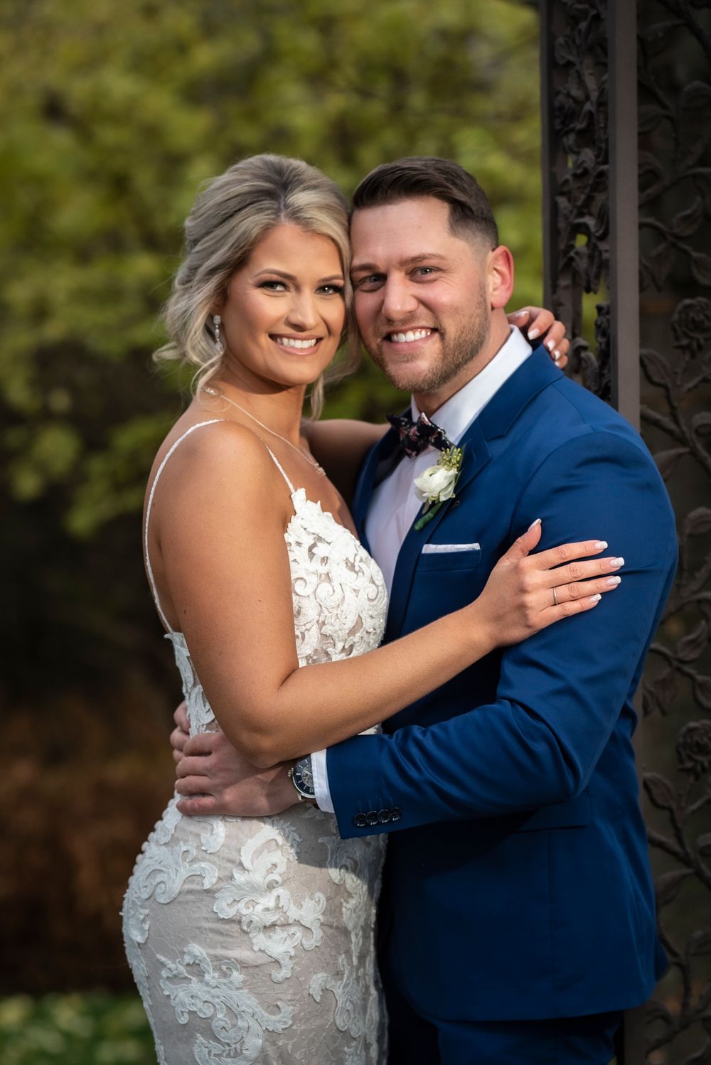 A newlywed couple embraces outdoors in wedding attire with the bride in white lace and groom in navy blue suit.