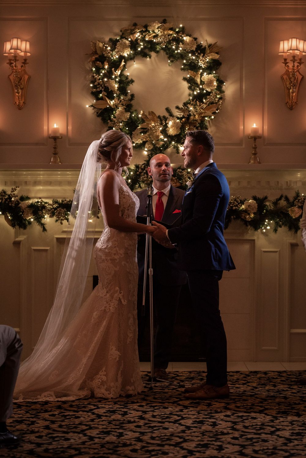 A wedding ceremony takes place in an elegant ballroom decorated with Christmas trees and warm lighting.