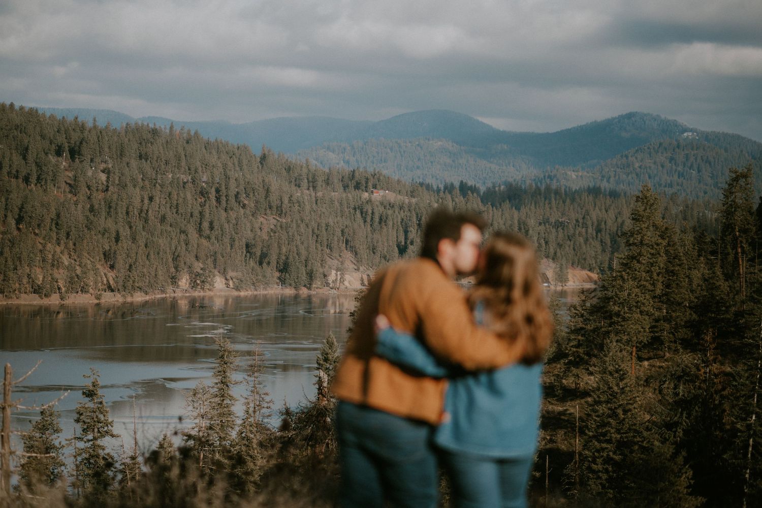 A couple embracing in front of a scenic lake surrounded by forested mountains.