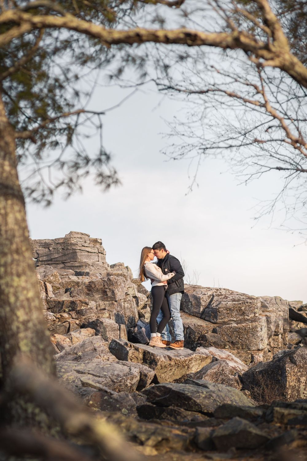 Couple embracing on rocky cliffs with tree branches framing the scenic outdoor setting.