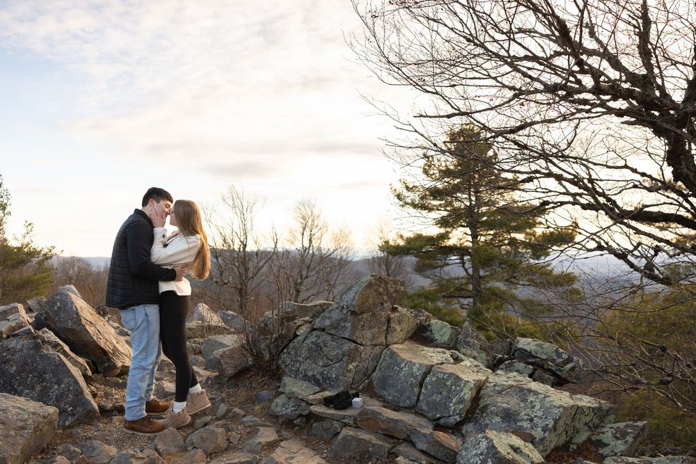 A couple shares a romantic moment at sunset on a rocky mountain overlook with scenic views in the background.