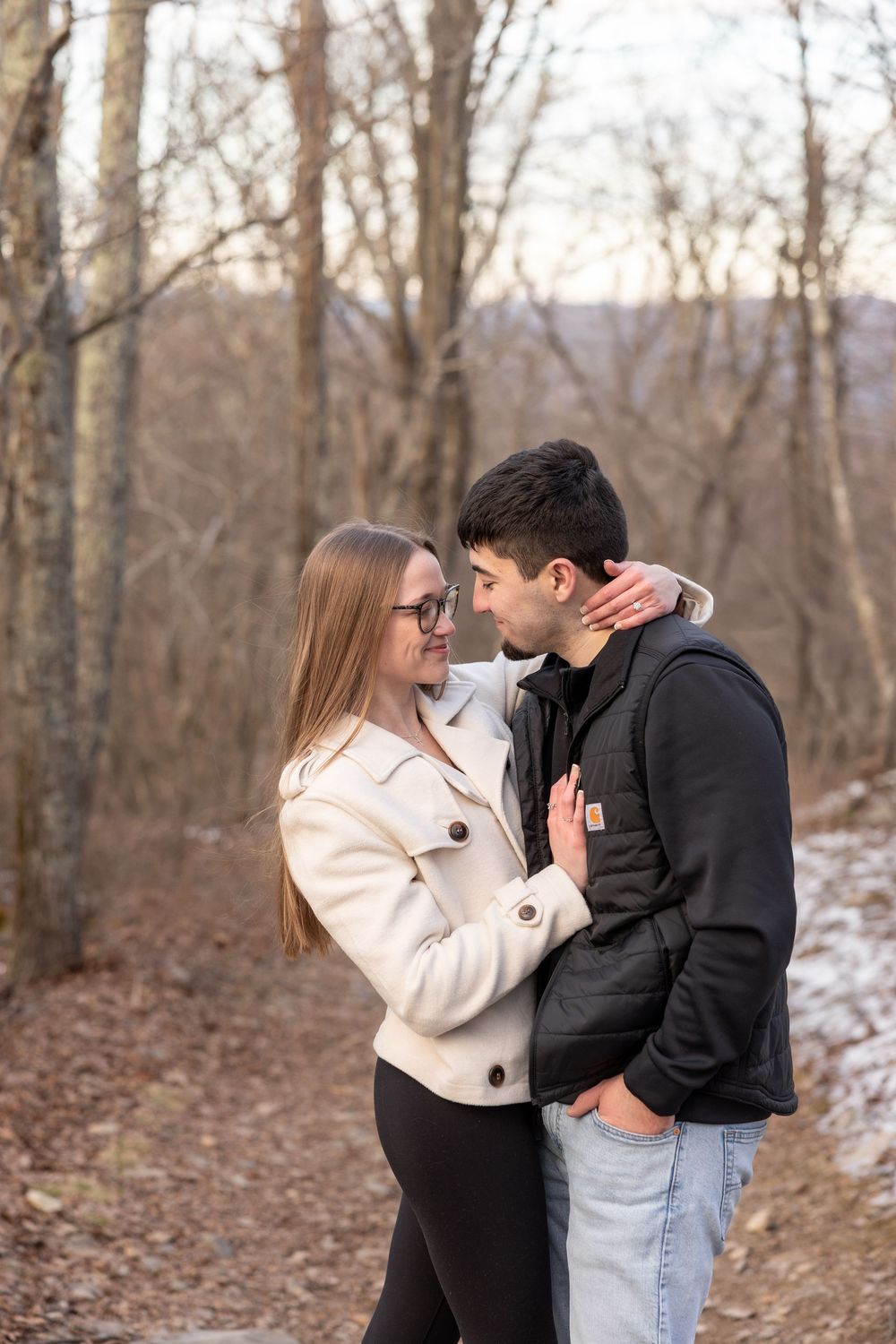 A couple embraces on a winter trail surrounded by bare trees and fallen leaves.