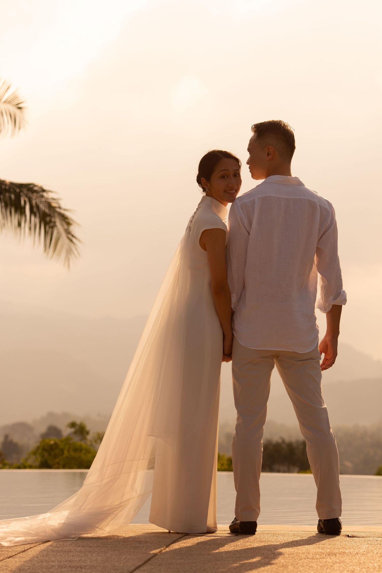 Bride and groom standing together at sunset overlooking Koh Samui, with soft golden light and flowing veil at a destination wedding.