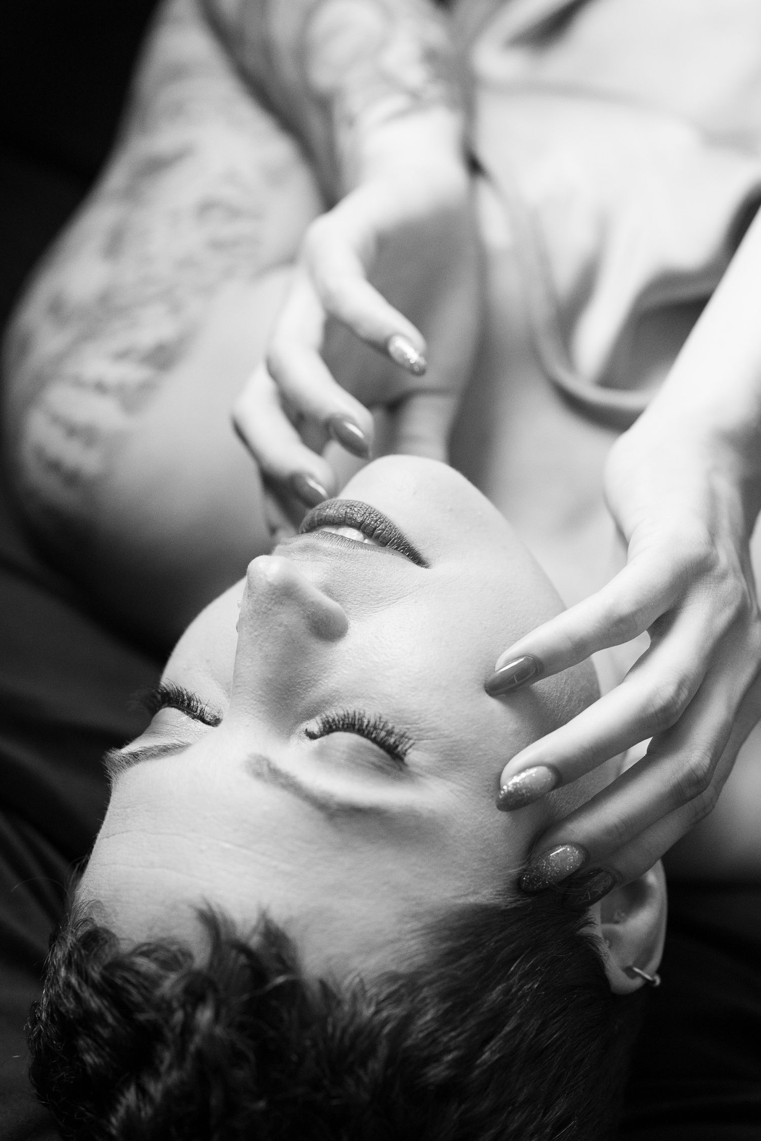 Black and white intimate portrait of a tattooed woman lying back with hands near her face.