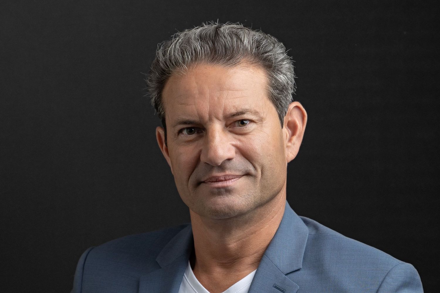 Professional headshot of man with gray hair wearing blue blazer against dark background.