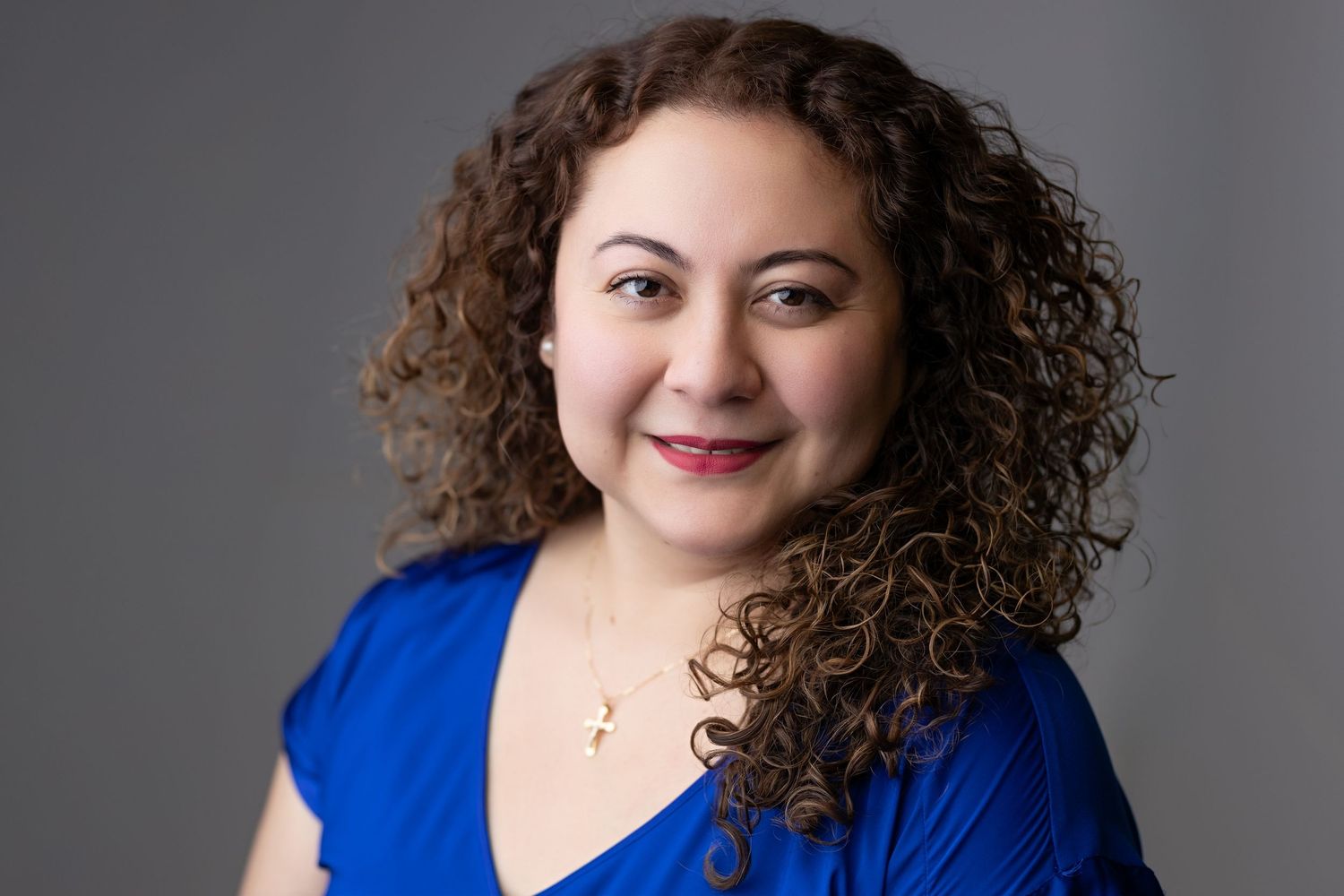Professional headshot of smiling woman with curly hair in blue top wearing gold cross necklace.