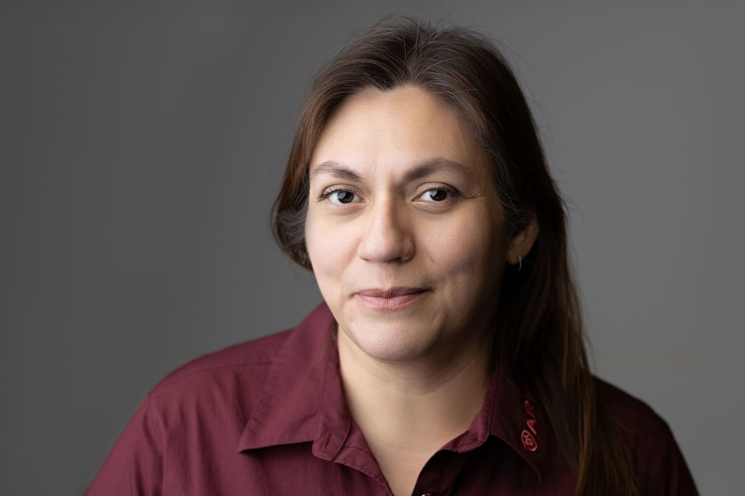 Professional headshot of woman with straight brown hair wearing burgundy shirt on gray background.