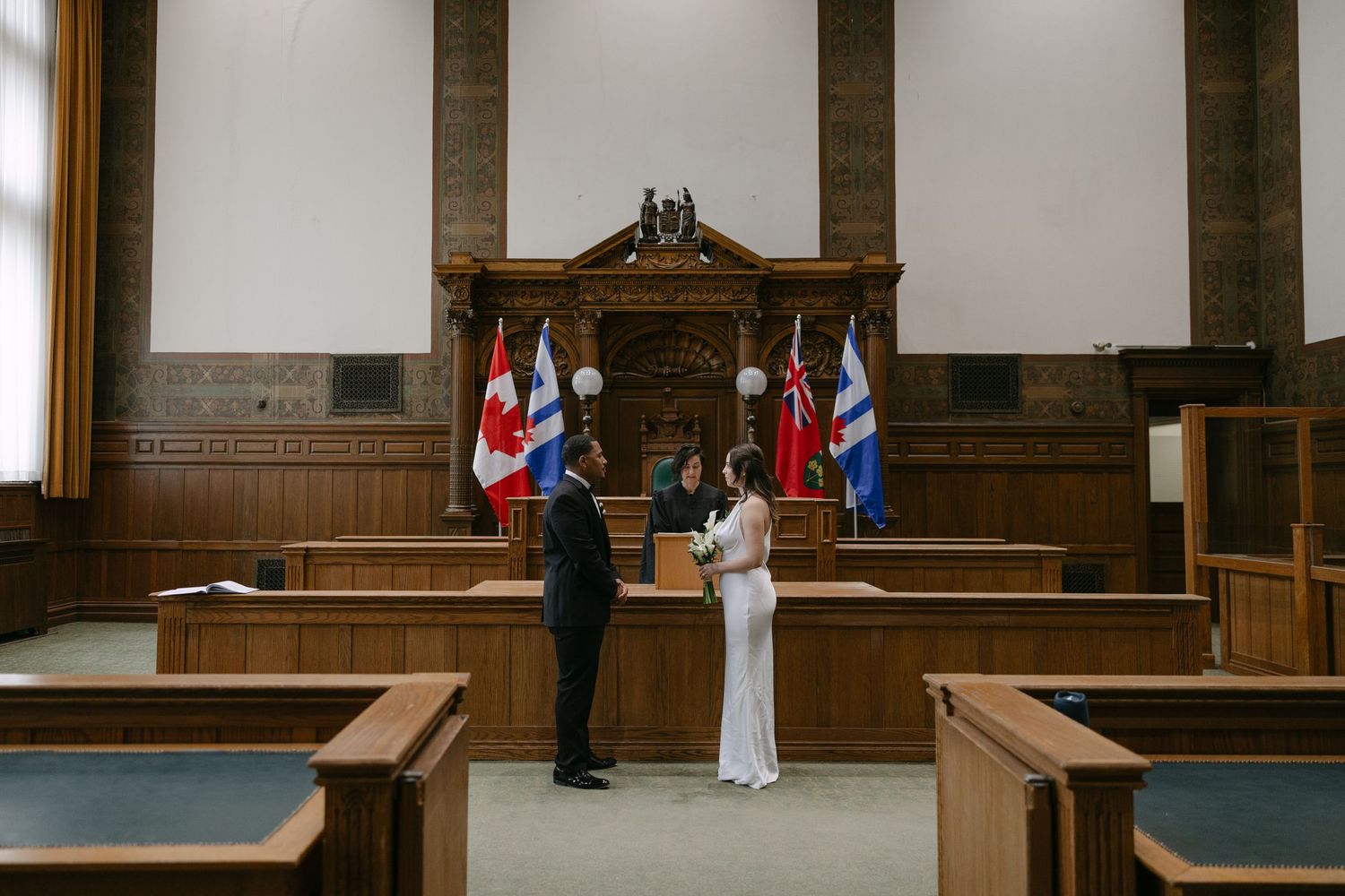 Wedding ceremony in progress at a historic courthouse with flags and wooden furnishings.The old city hall Toronto wedding photographer Daria