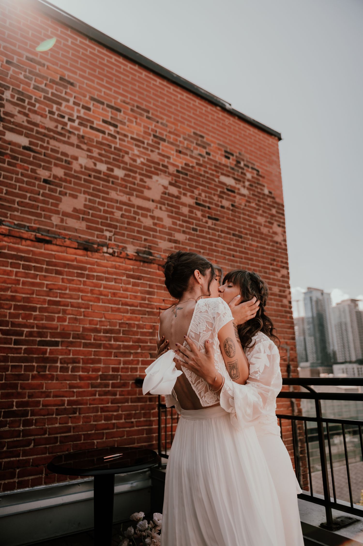 Two people in white wedding dresses share an intimate moment against a brick wall backdrop in an urban setting.