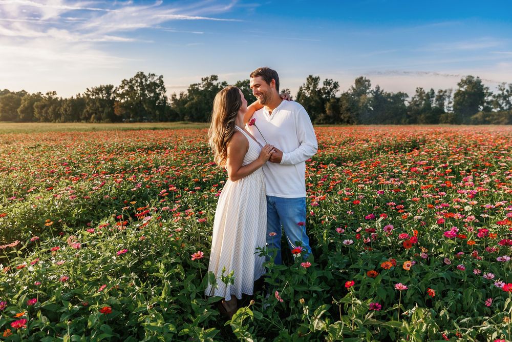 A couple shares a romantic moment in a vibrant field of red and pink zinnias during a sunny summer day.