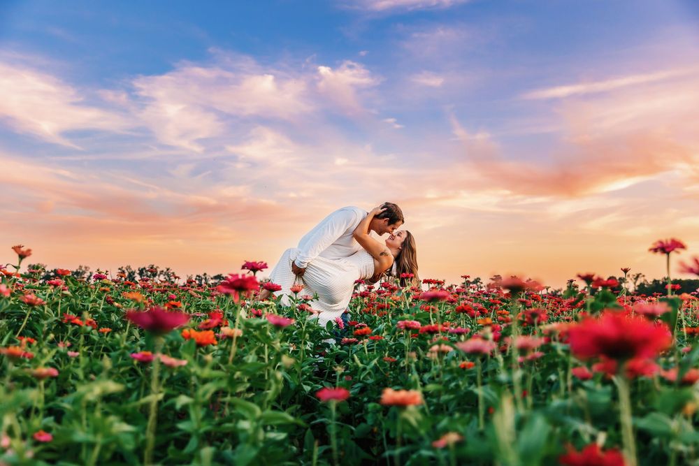 A couple embraces in a vibrant red flower field during a colorful sunset with dramatic clouds in the sky.