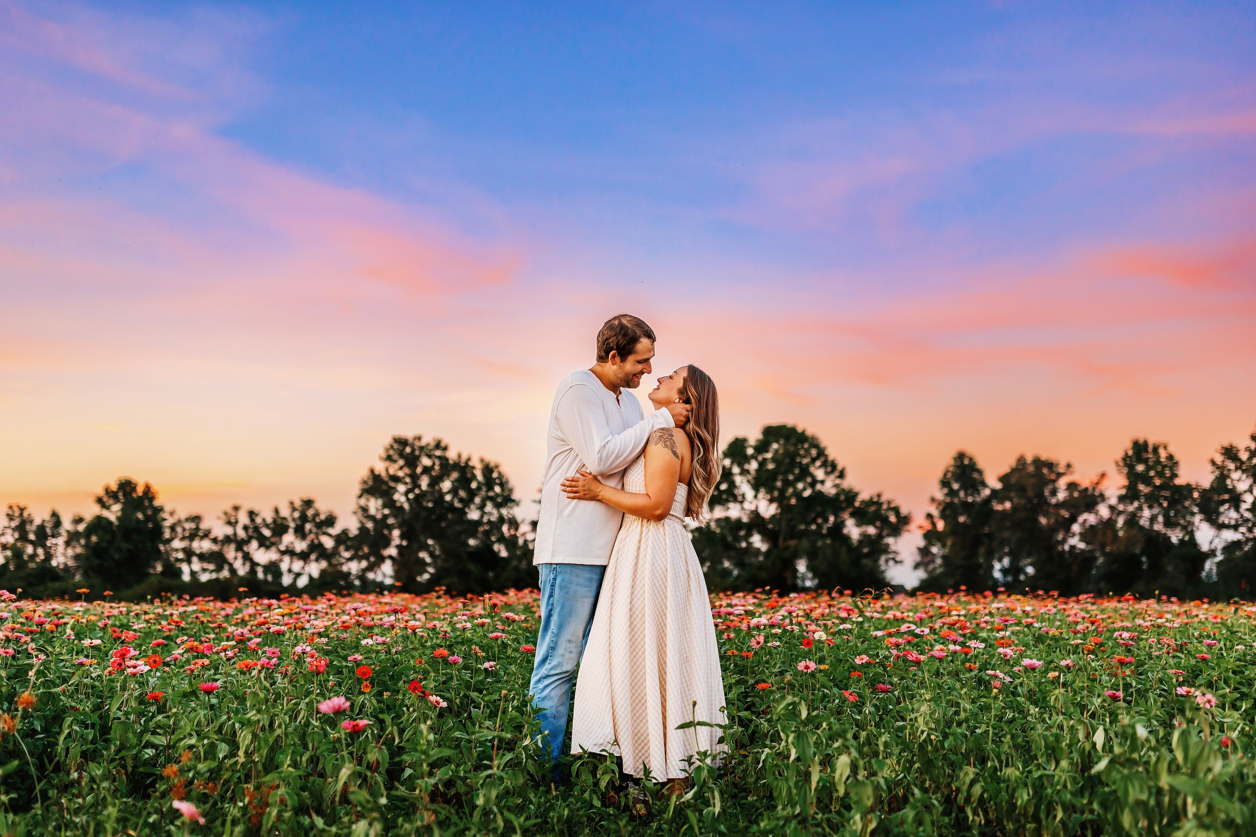 A romantic couple embraces in a vibrant poppy field at sunset with pink and purple skies overhead.