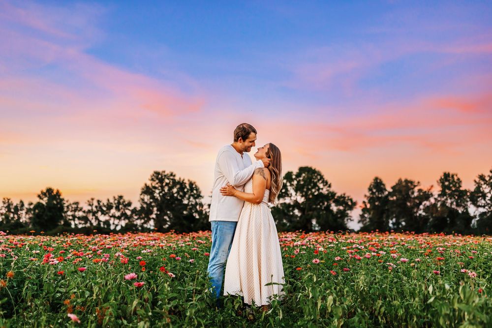 A romantic couple embraces in a vibrant poppy field at sunset with pink and purple skies overhead.