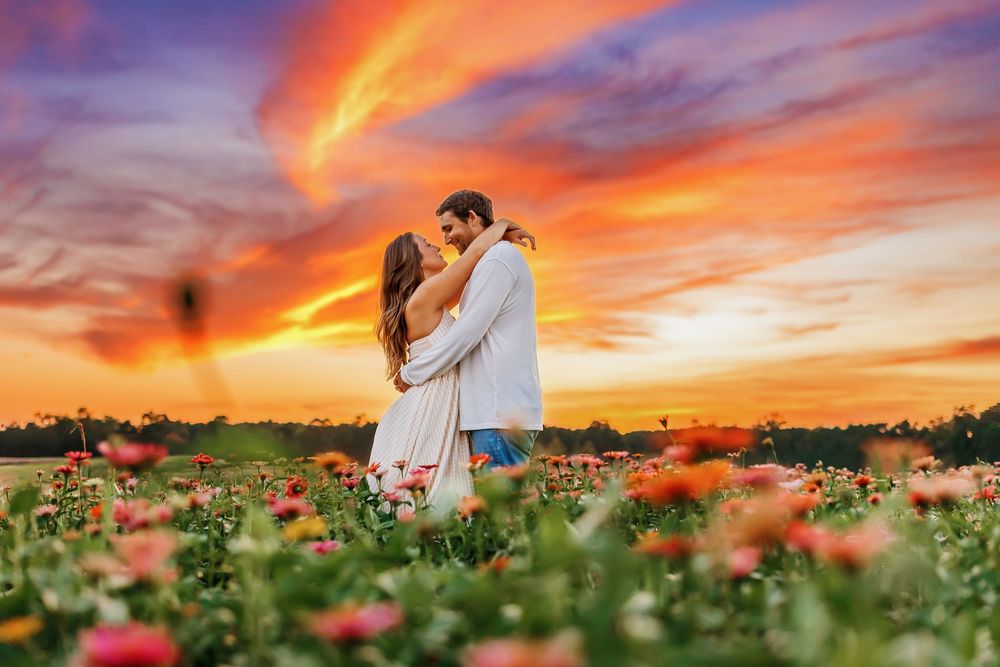 Romantic couple embraces in a vibrant poppy field at sunset with dramatic orange and purple sky overhead.